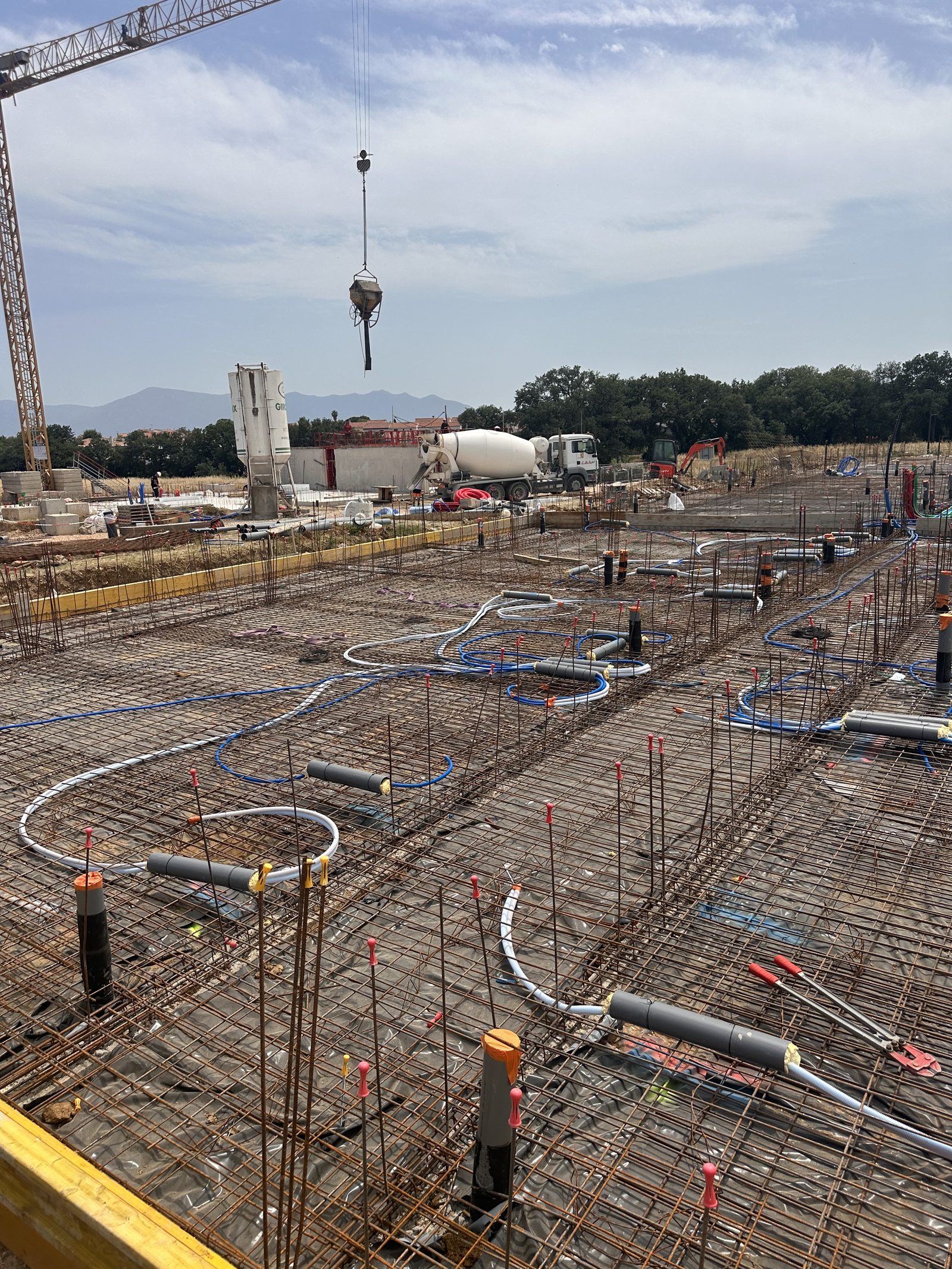 Dalle de résidence ferraillée avant coulage, avec réseaux PER, fourreaux en attente et réservations, grue avec benne à béton et camion toupie en fond